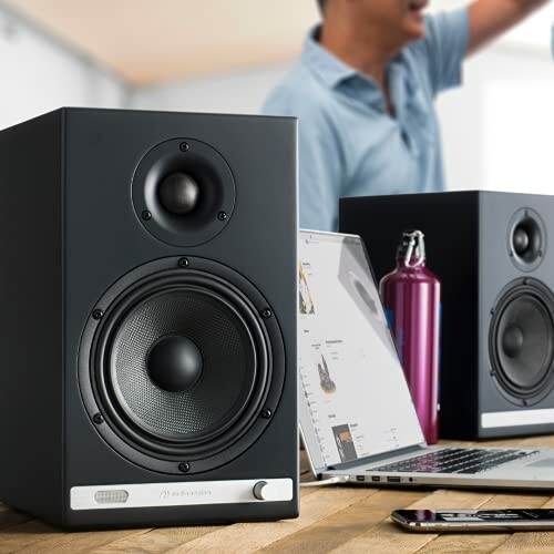 Speakers on a desk with a laptop and a person in the background.
